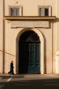 A lone person in black walks by a large ornate door on a sunny day.