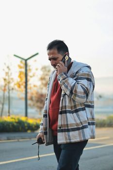 Casual man in plaid jacket walks outdoors talking on smartphone in an autumn setting.