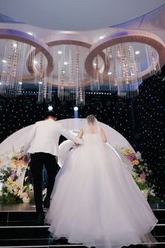 Bride and groom walking towards floral arch under chandeliers in elegant indoor wedding.