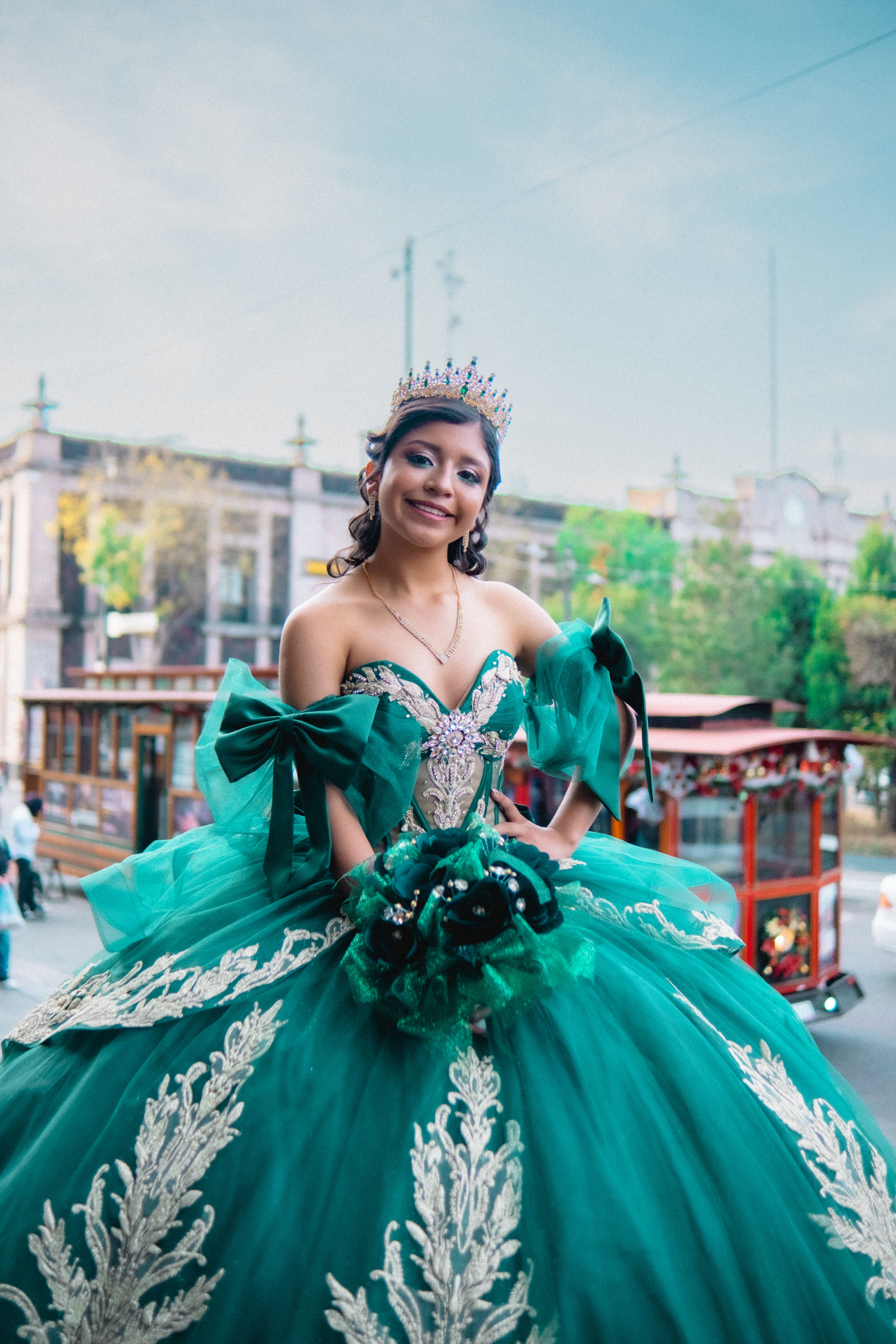 Young woman in green quinceañera dress with tiara stands outdoors, smiling.