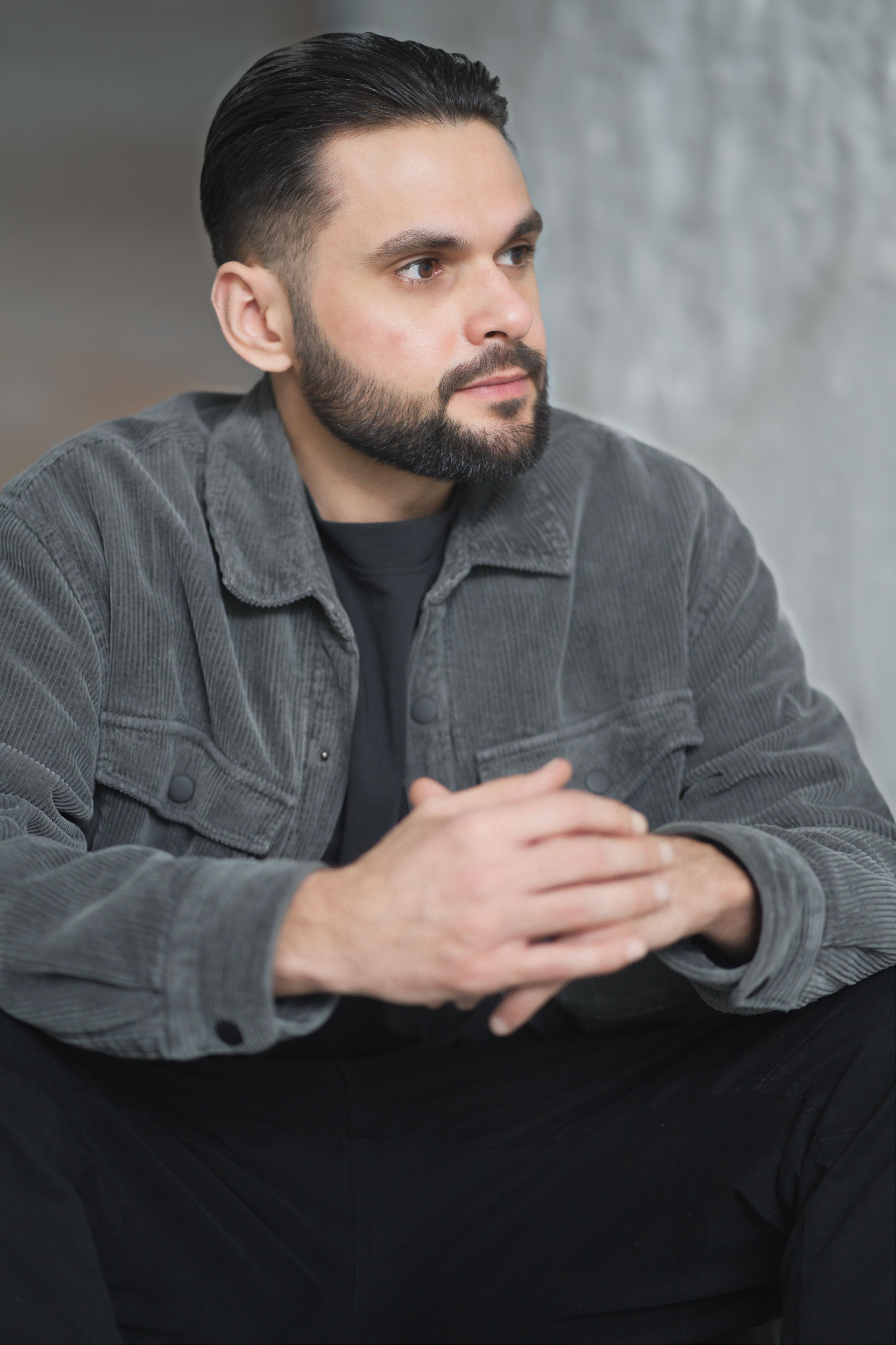 Thoughtful adult man with beard seated indoors wearing a gray jacket.