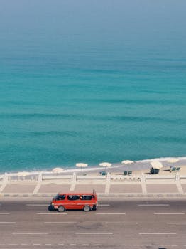 Red van travels along a serene coastal highway with turquoise waters and beach umbrellas.