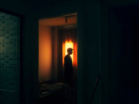 Silhouette of a man standing by a bright window in a dimly lit room, creating a moody and introspective scene.