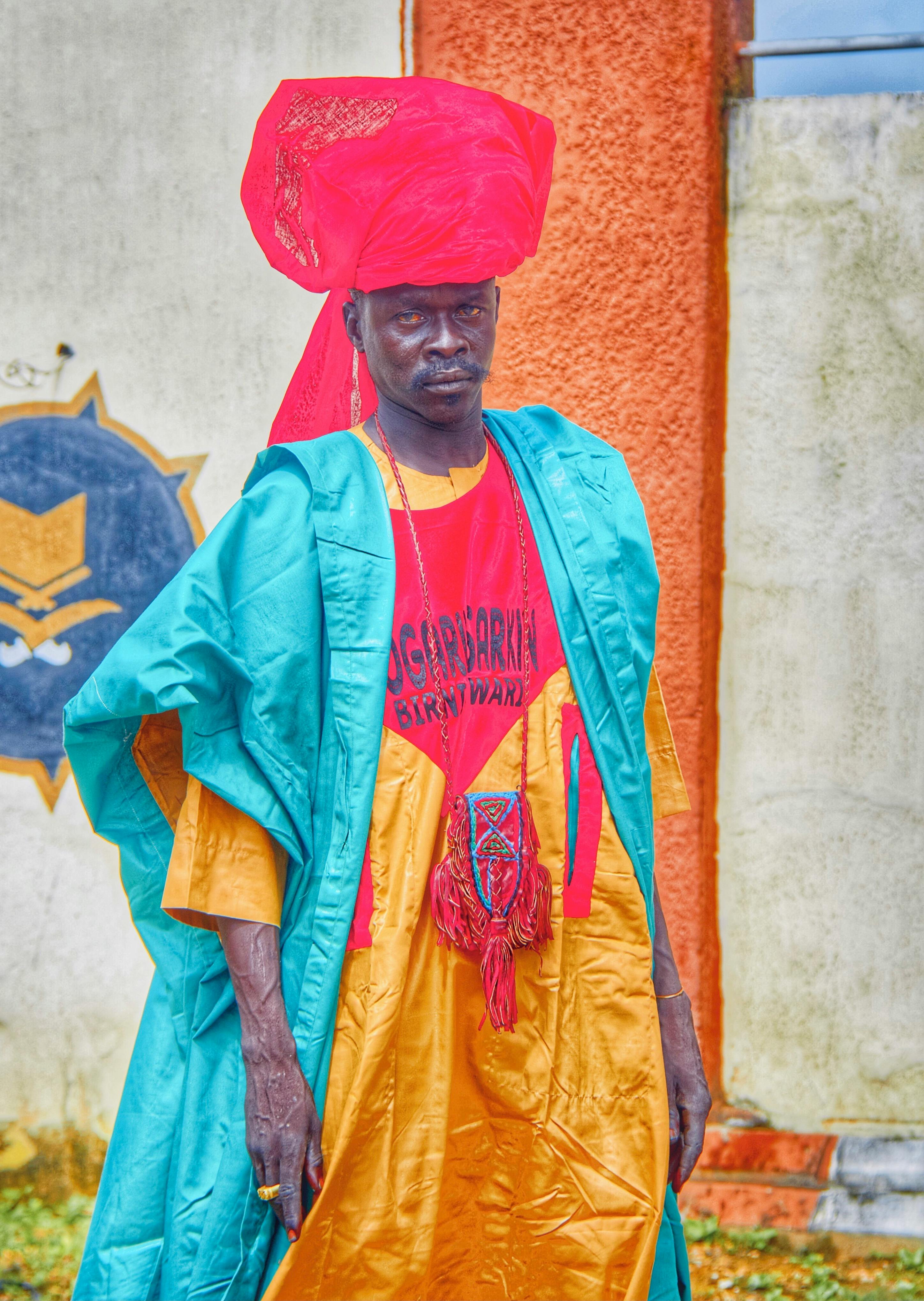 Middle Eastern man in colorful traditional attire with turban, standing outdoors against a textured wall.