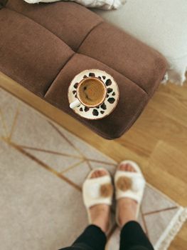 Top view of coffee on a brown sofa with person in slippers on a rug.