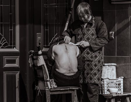Monochrome image of a street barber giving an outdoor haircut to a seated individual, displaying craftsmanship.