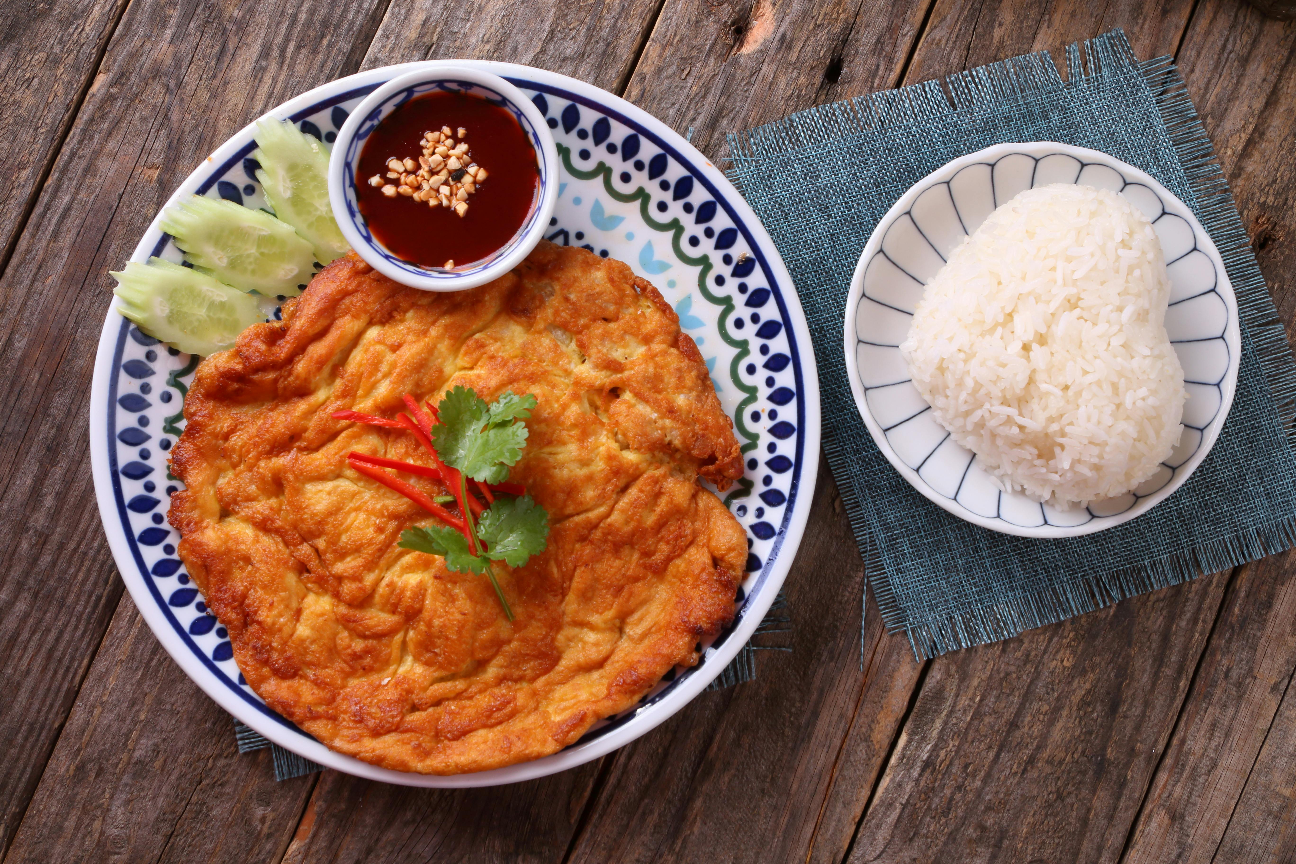 Delicious Thai omelette with rice and chili sauce garnished with cilantro and cucumbers.