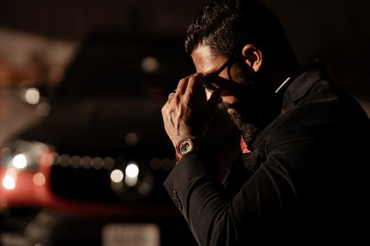 A man in a coat adjusts his sunglasses by a car in a dimly lit area.
