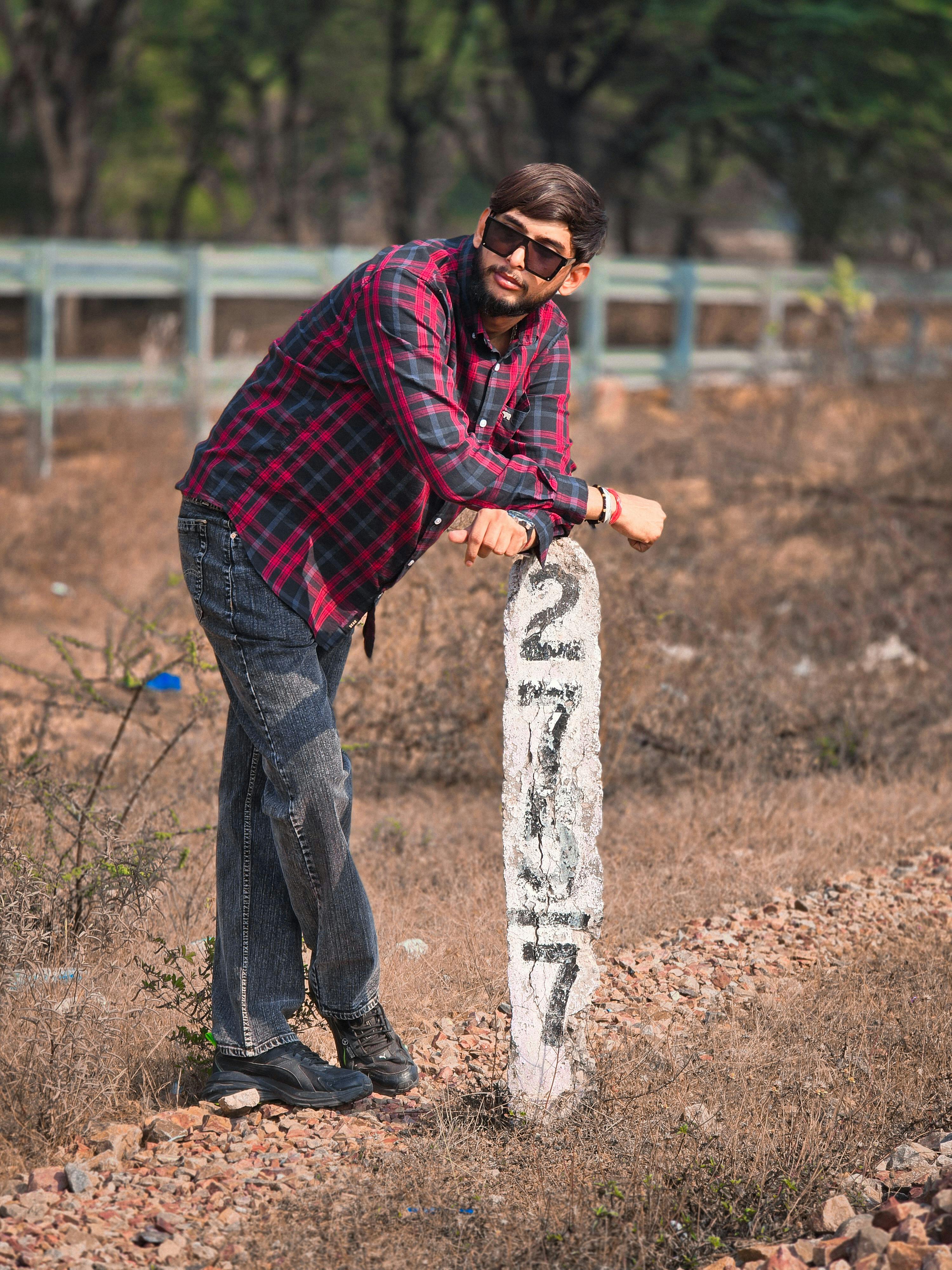Man in plaid shirt leaning on a mile marker in a rural outdoor setting, conveying a relaxed vibe.