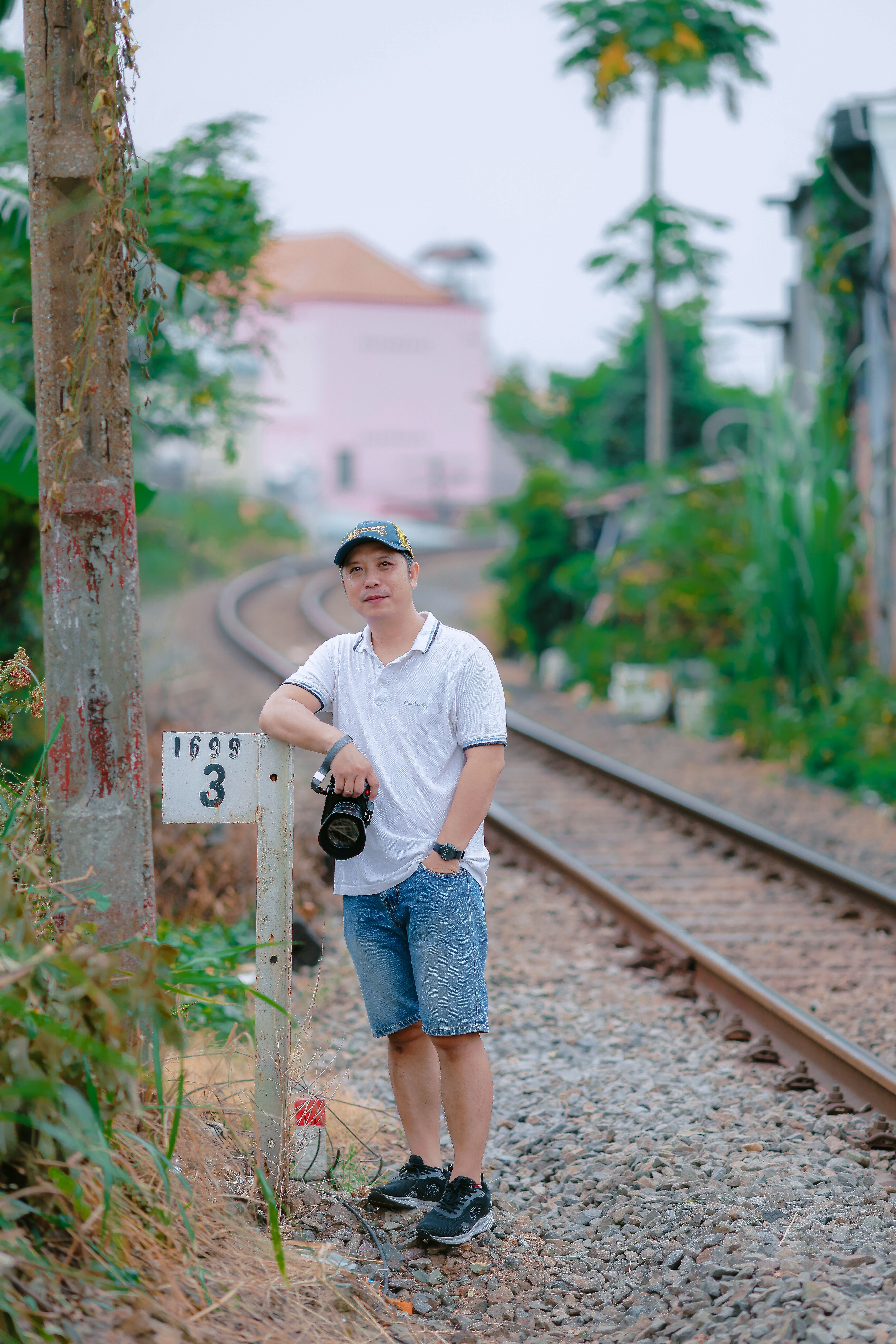 Man in casual outfit holding camera beside railroad tracks in a rural setting.