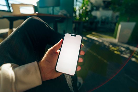 Close-up of a hand holding a smartphone with a blank screen in a sunlit modern office environment.