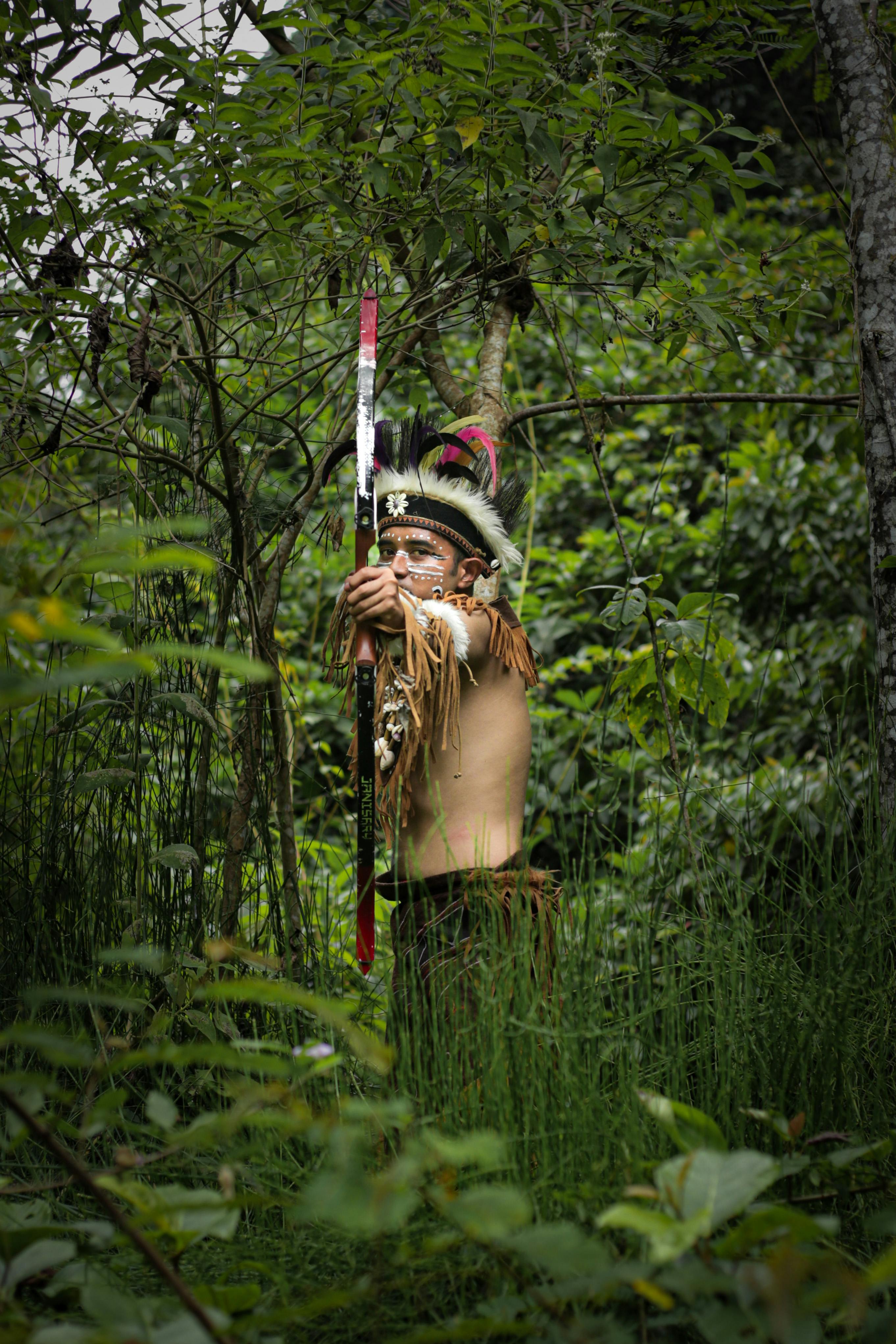 Indigenous archer stands in a lush jungle, showcasing traditional attire and bow, amidst vibrant greenery.