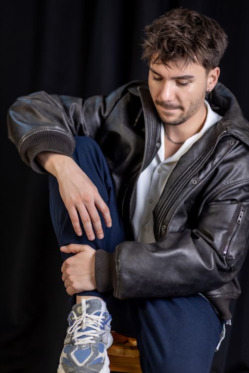 Free Young man wearing a leather jacket and sneakers sitting against a black background. Stock Photo