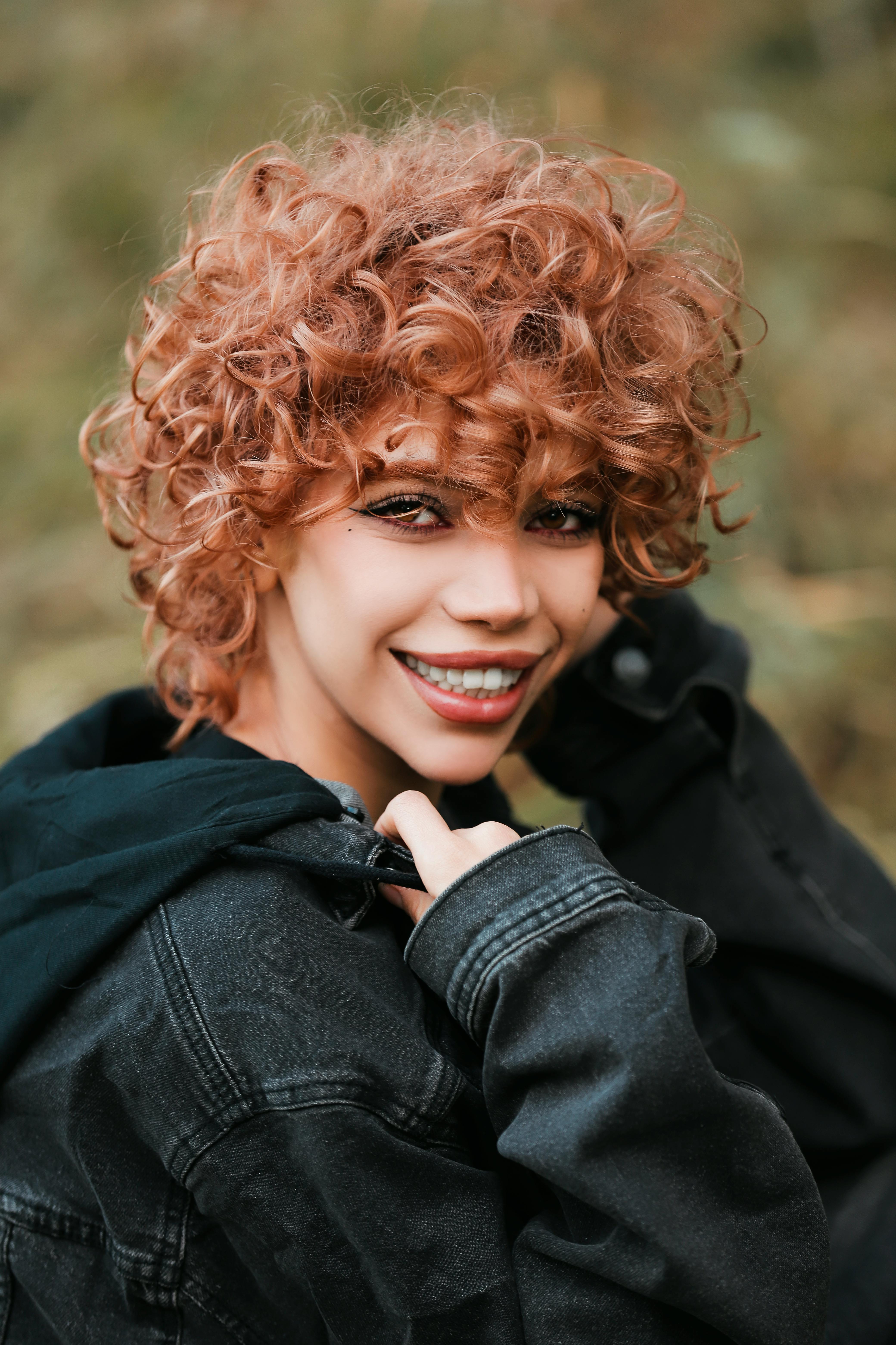 Portrait of a woman with curly hair wearing a jacket, smiling outdoors.