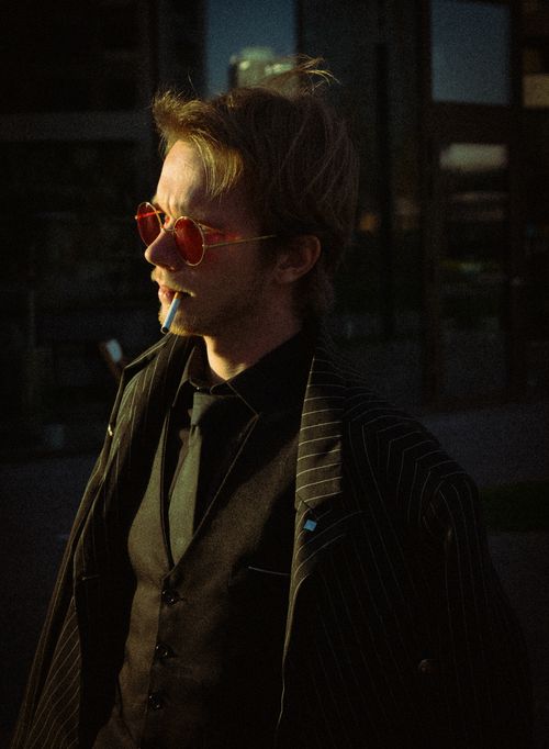 Free A fashionable man in pinstripe suit and red glasses with cigarette, captured outdoors at dusk. Stock Photo