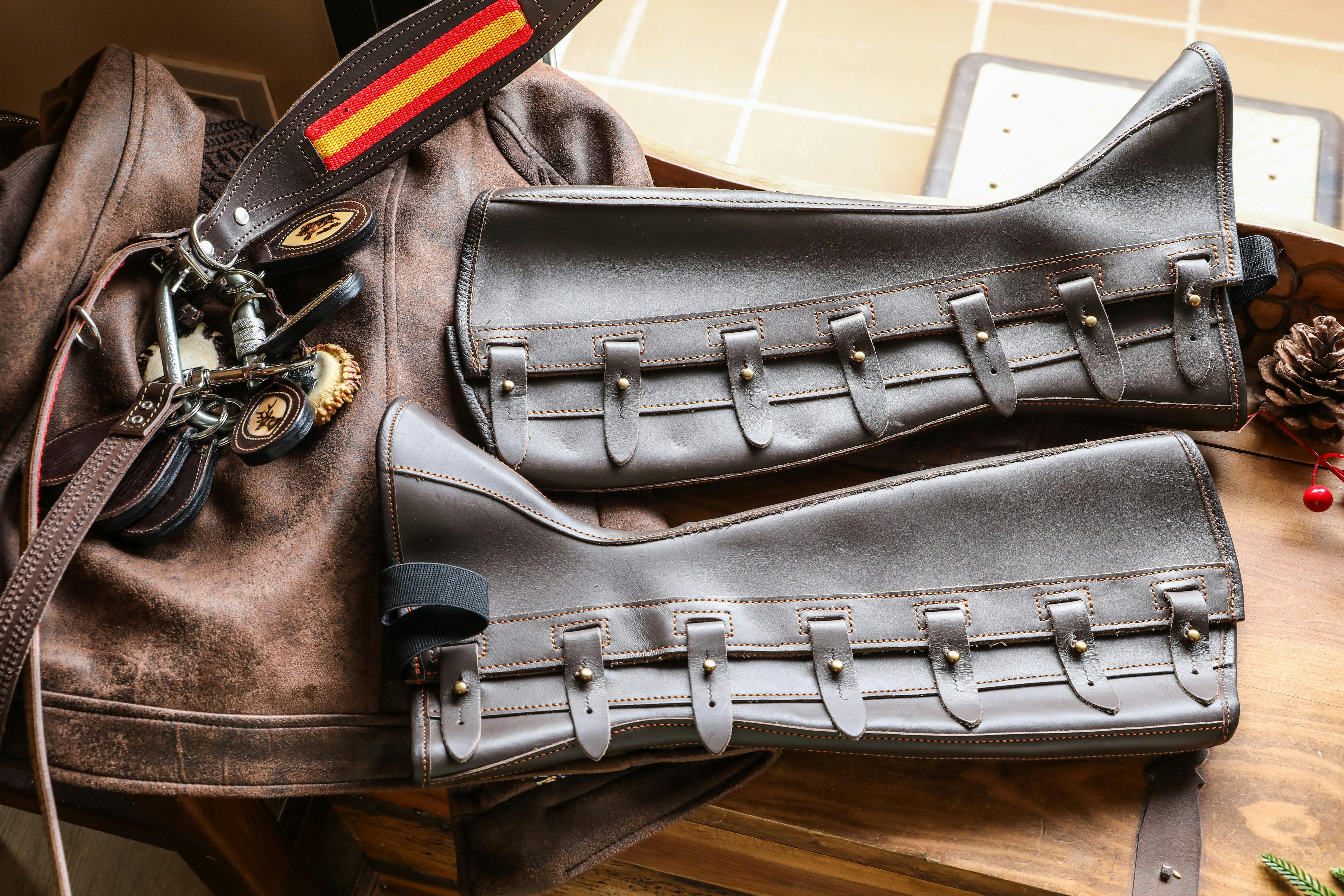 Close-up of leather equestrian gear including gaiters and saddle accessories on a wooden table.