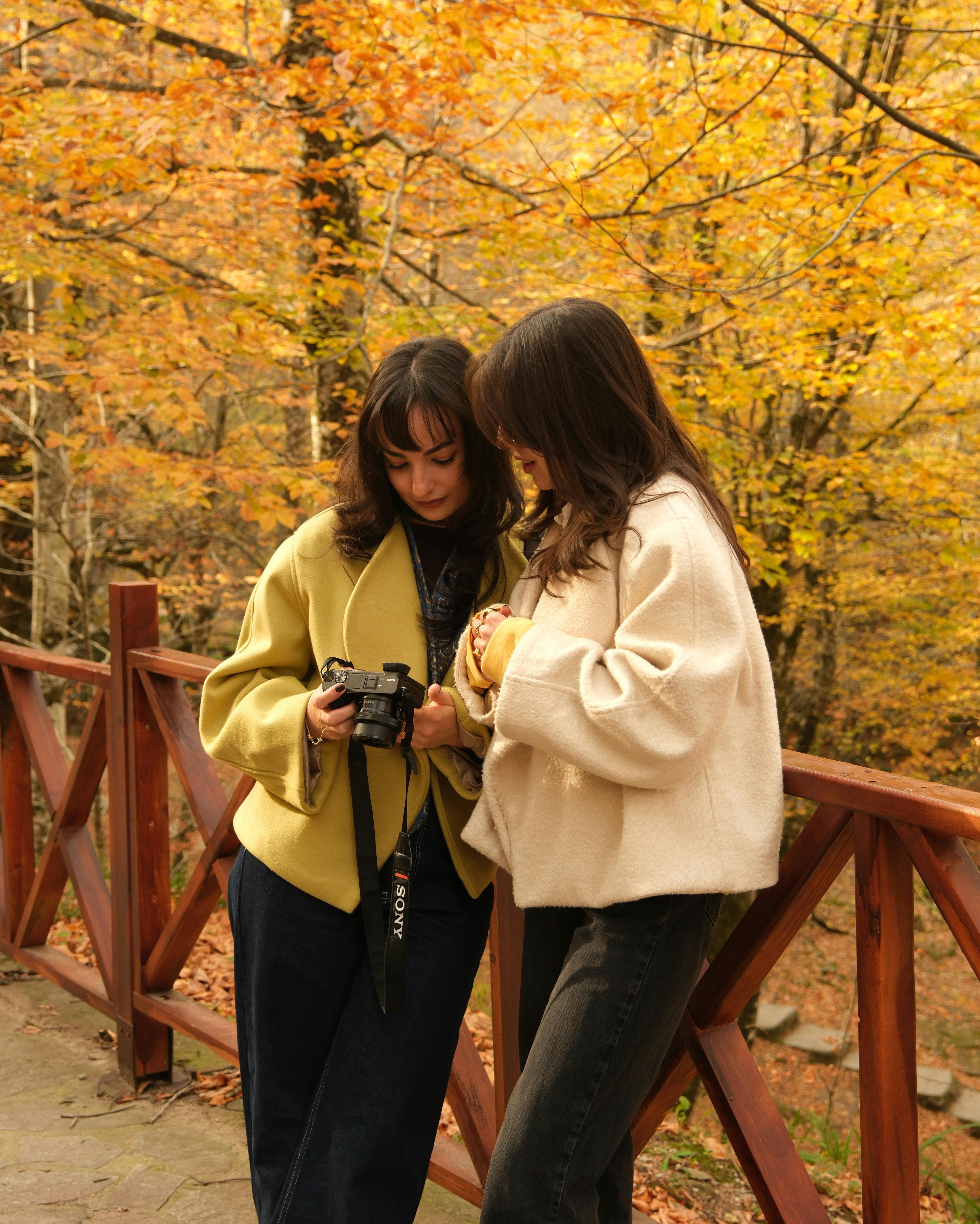 Two women review photos on a digital camera in a vibrant autumn forest.