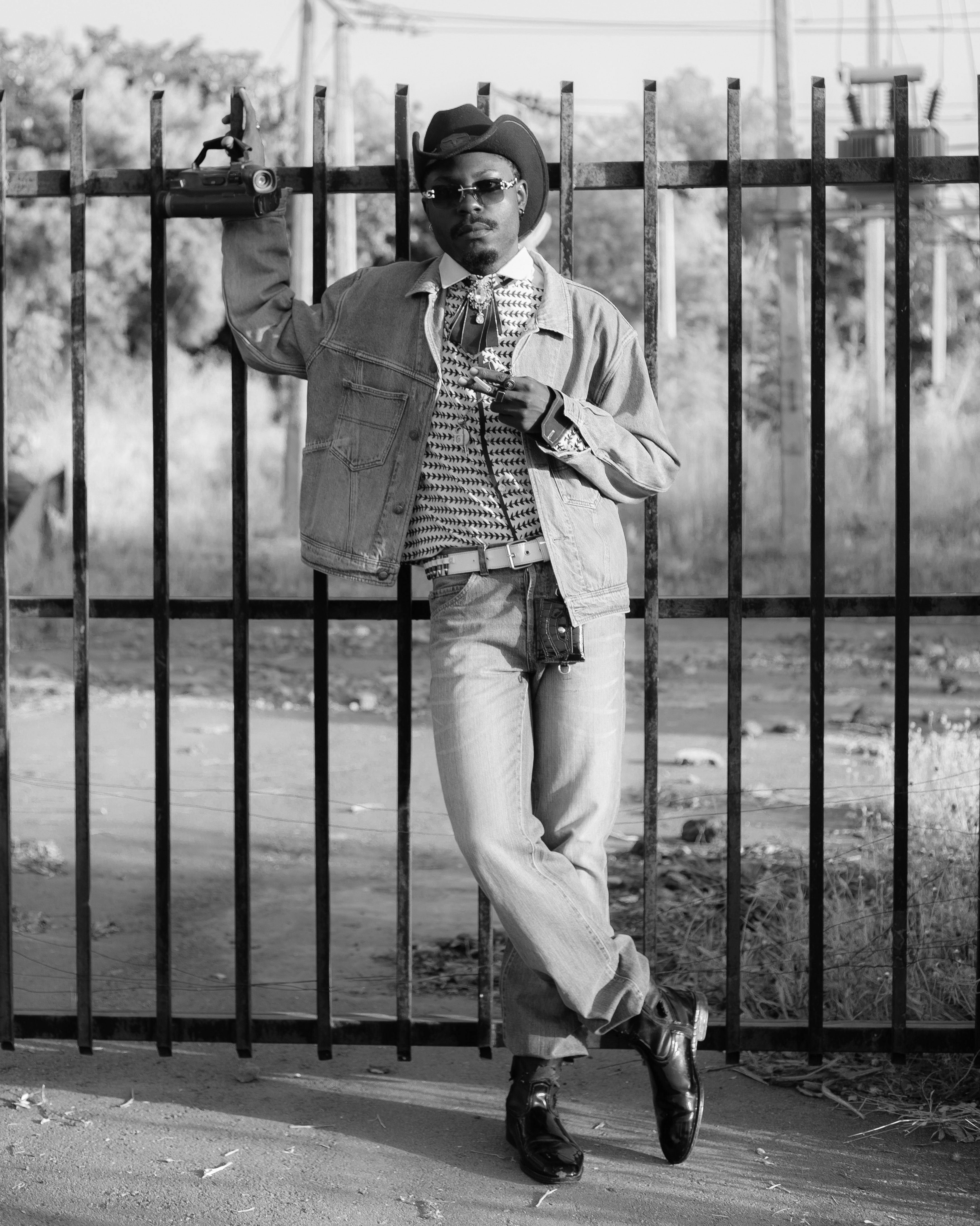 A fashionable man in Western attire poses confidently against a metal gate.