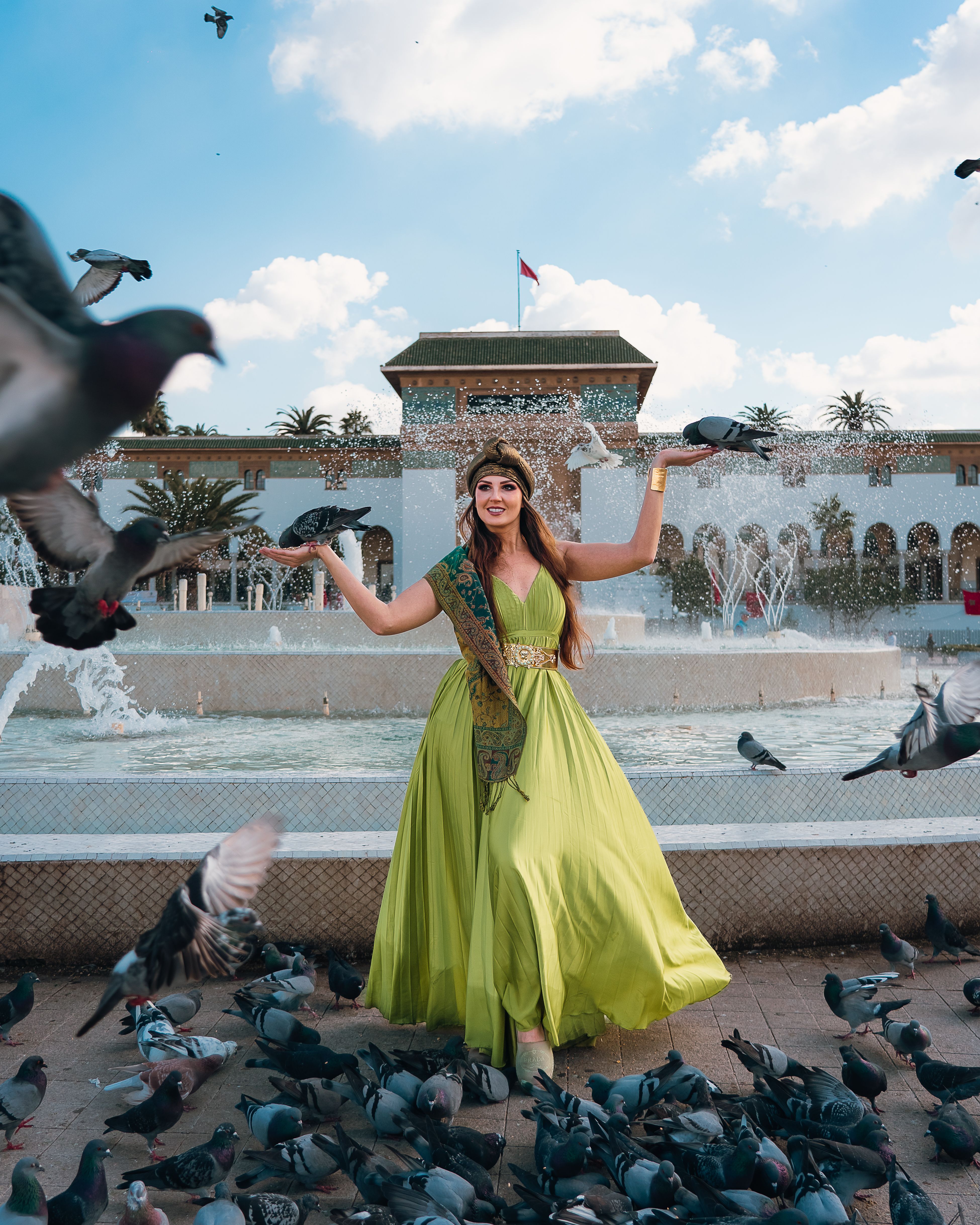 Woman in green dress surrounded by pigeons near a fountain on a sunny day.