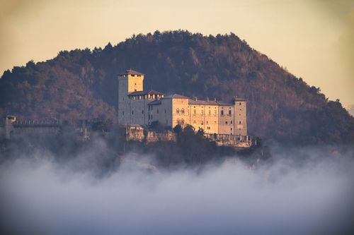 Free A serene view of a historic castle emerging from the mist against a wooded hill at dawn. Stock Photo