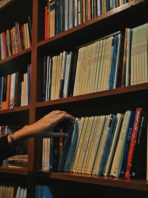 Free A hand reaches for a book on a wooden bookcase in a quiet, dimly lit library. Stock Photo