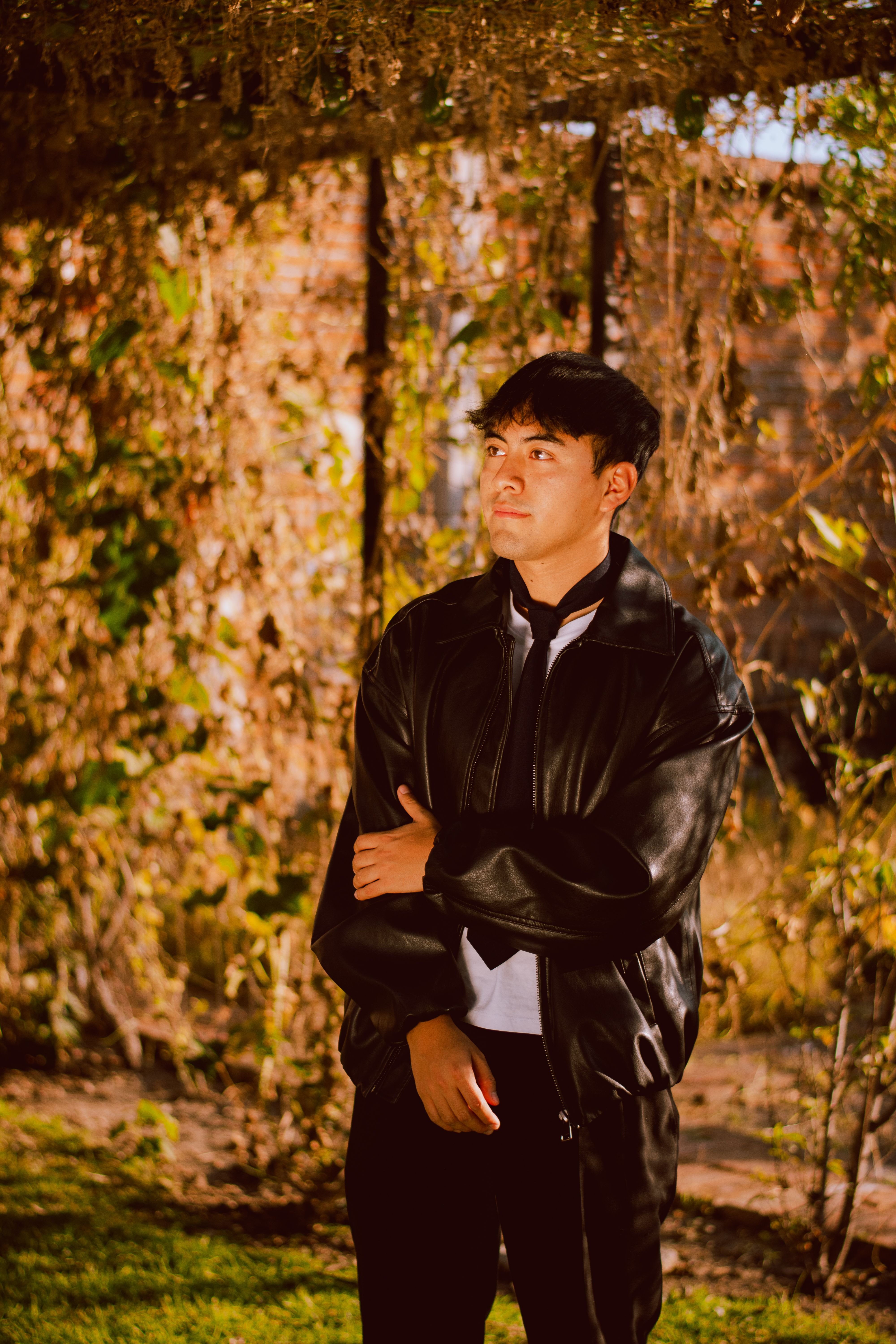 A young man stands confidently in an autumn garden wearing a stylish black leather jacket.