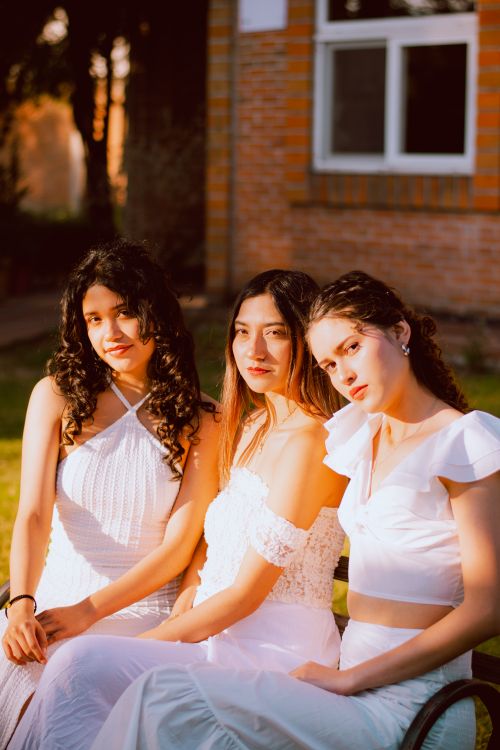 Free Three young women in white dresses pose gracefully in warm sunlight outdoors. Stock Photo
