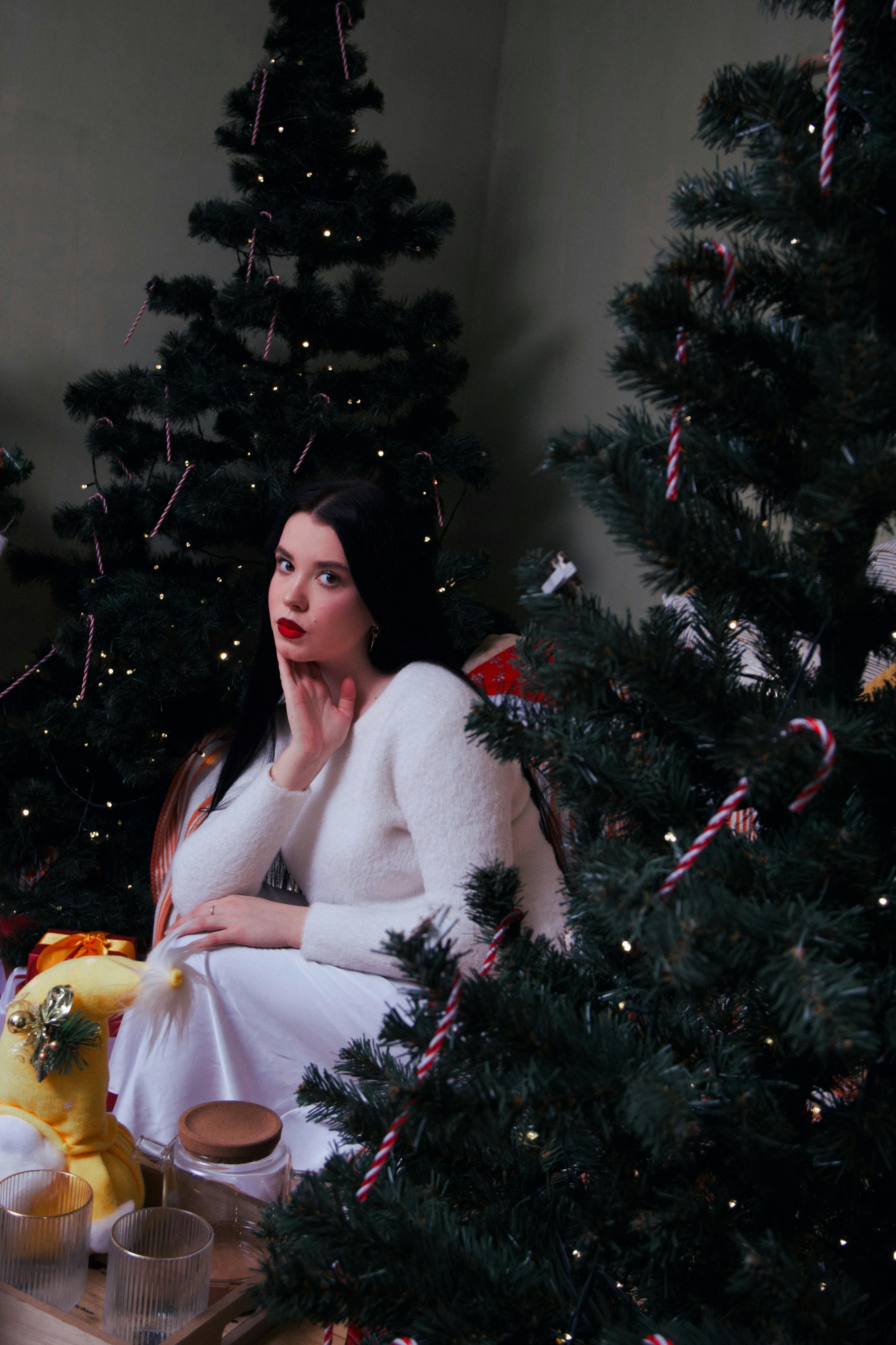 Woman in winter white attire sitting among decorated Christmas trees indoors.