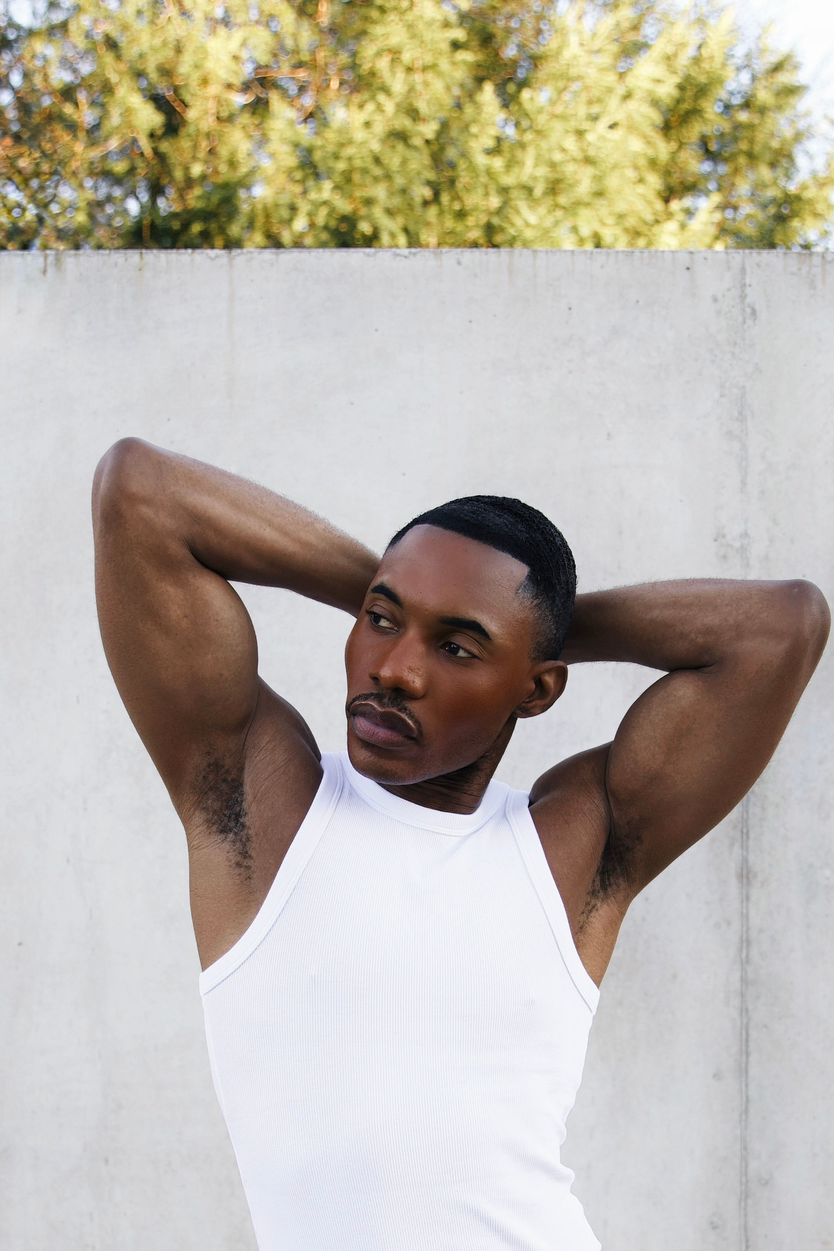 A confident man in a white tank top posing outdoors in Austria with arms behind head.
