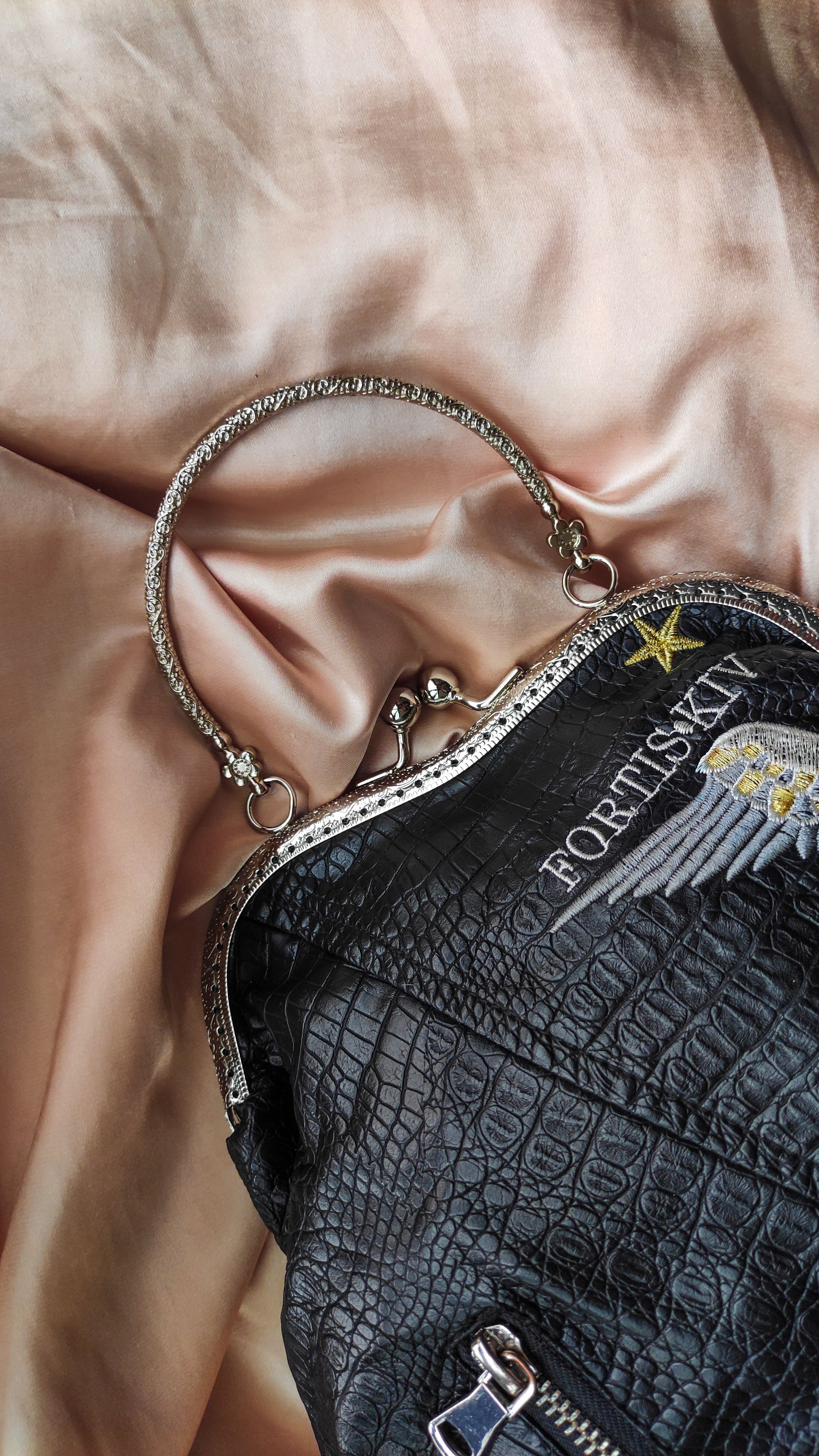Elegante bolsa de couro preto com detalhes metálicos sobre fundo de seda rosa suave.