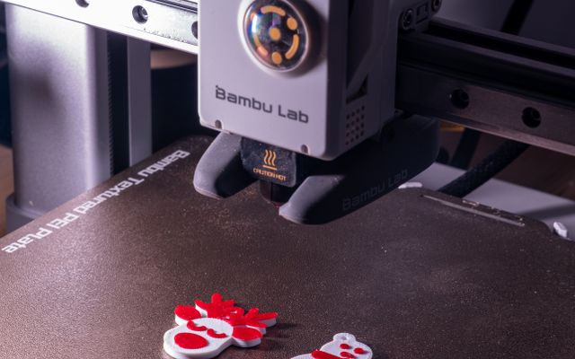 A Bambu Lab print head with a round fan over a textured build plate holding freshly printed red and white snowman decorations