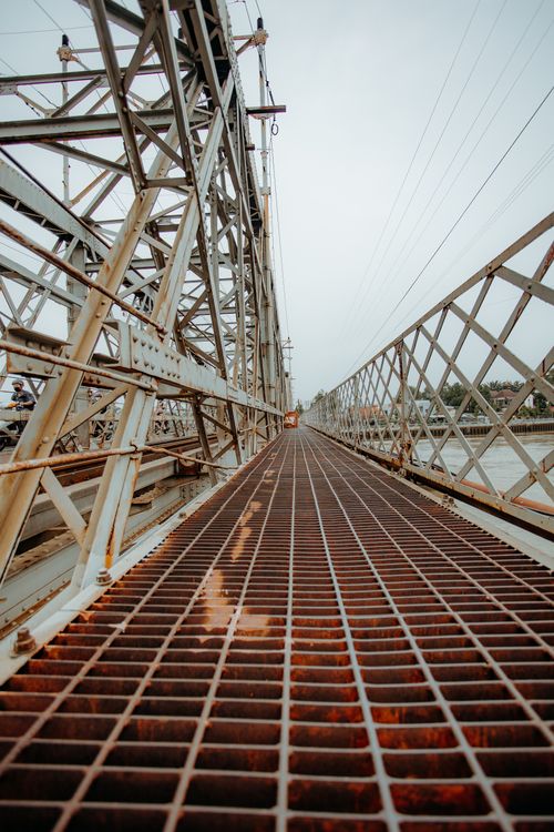 Free Perspective view of a rusty iron bridge stretching over a river under cloudy skies, showcasing industrial design. Stock Photo