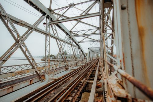Free A rusty steel railway bridge extends over a calm river, creating a dramatic perspective. Stock Photo