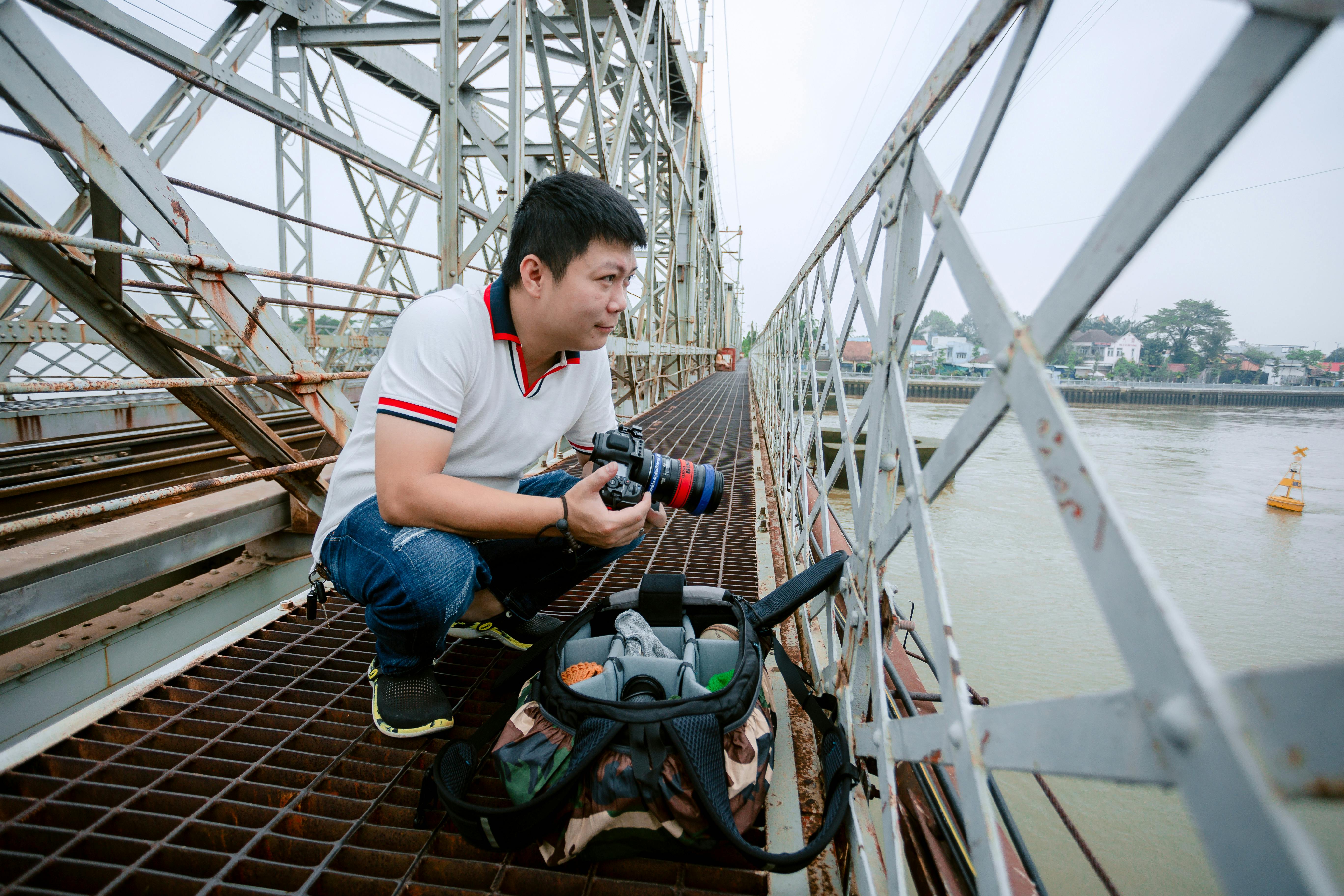 A man crouches on a bridge, photographing a river scene with professional camera gear.
