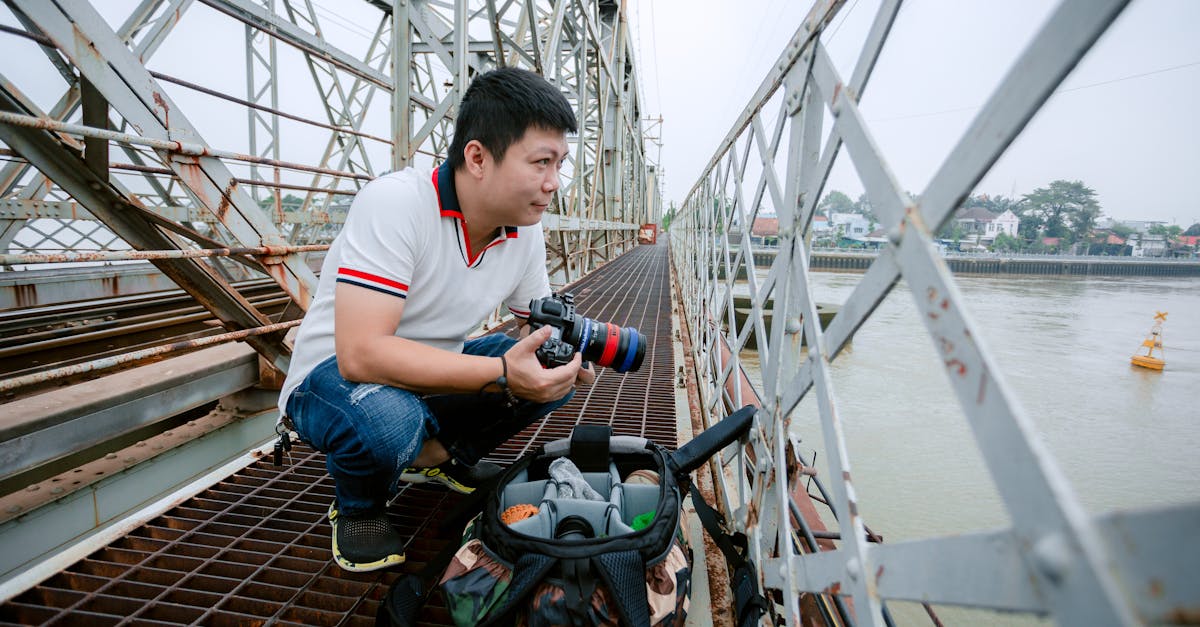 A man crouches on a bridge, photographing a river scene with professional camera gear.