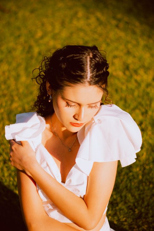 Free Beautiful woman with curled hair in a white dress, sunlit outdoors, serene expression. Stock Photo