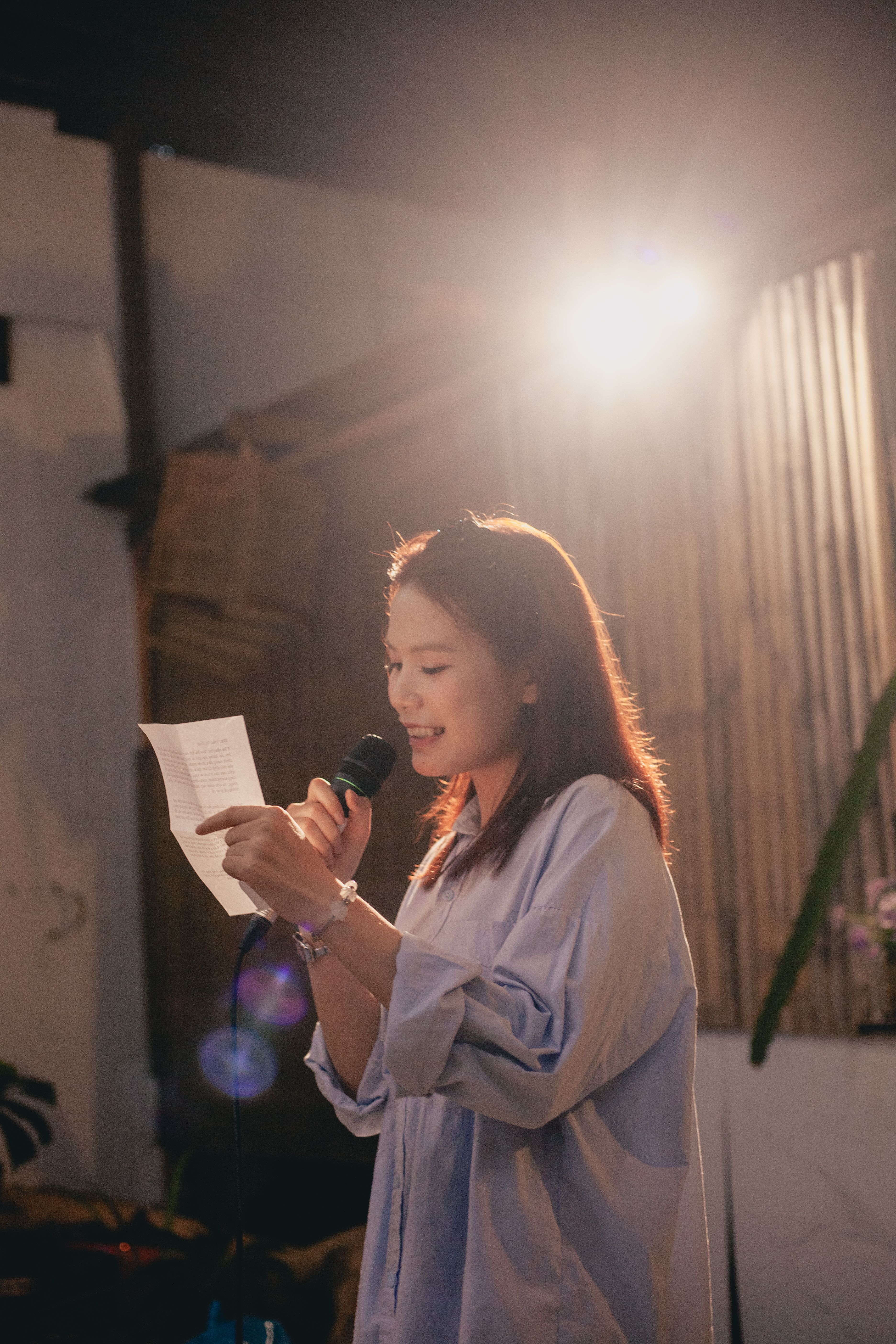 A woman delivers a speech or performance under warm lighting indoors with natural decor.