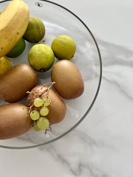 A glass bowl of fresh fruit including golden kiwis, grapes, limes, and a banana on a white marble surface.