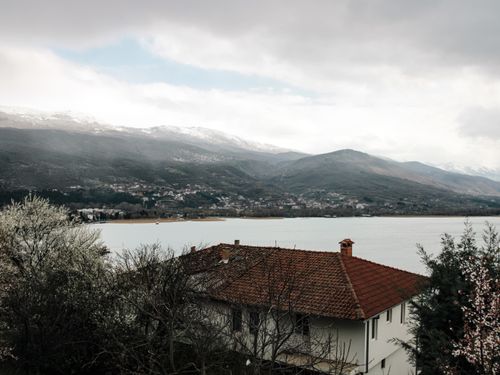 Free Tranquil lakeside scene with a house, mountains, and blossoming trees in early springtime. Stock Photo