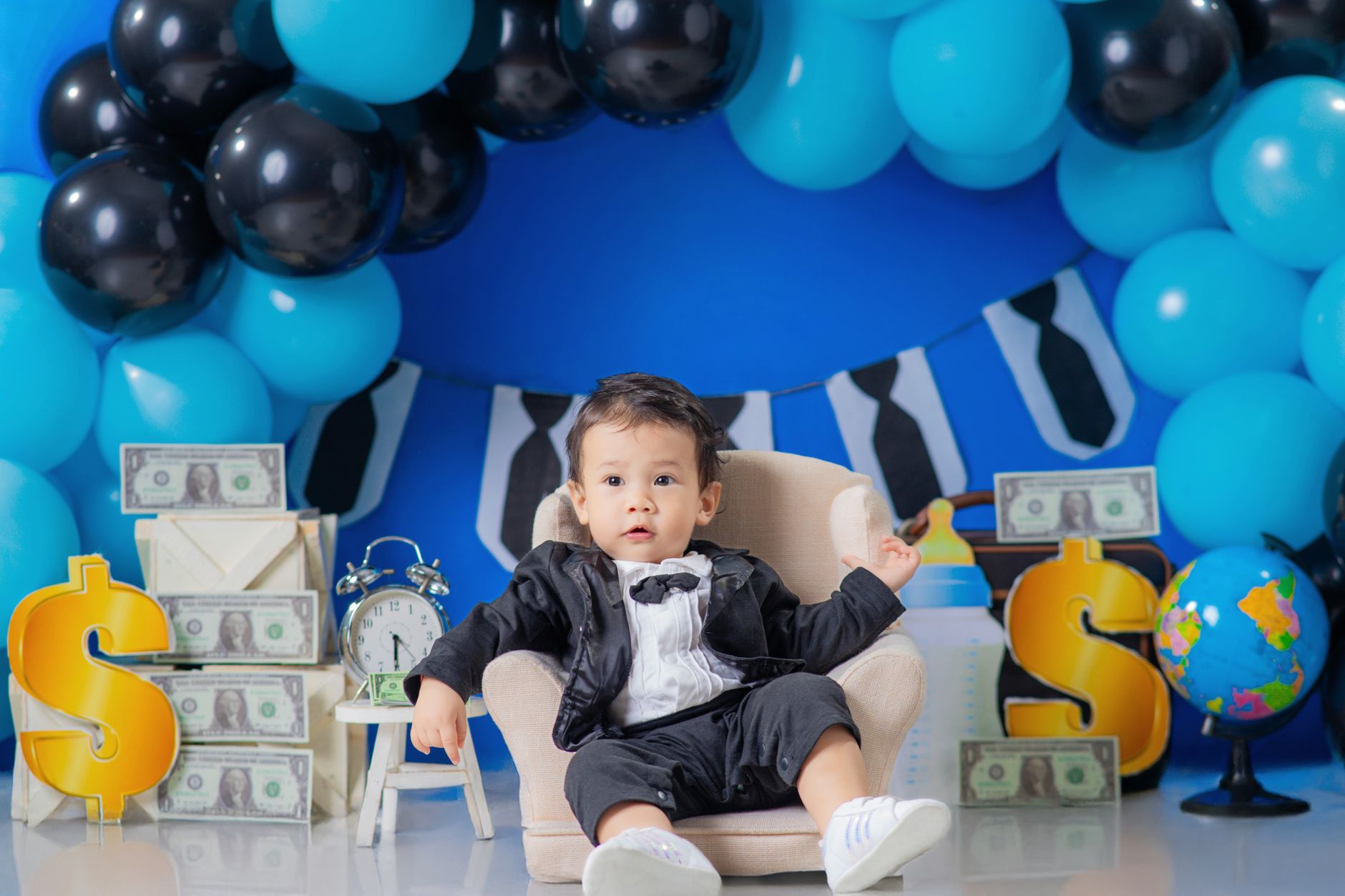 Charming photo of a baby in formal attire surrounded by dollar signs and blue balloons.