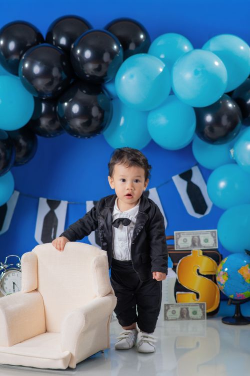 Free Charming baby in a tuxedo stands beside a chair, surrounded by festive balloons and decor. Stock Photo