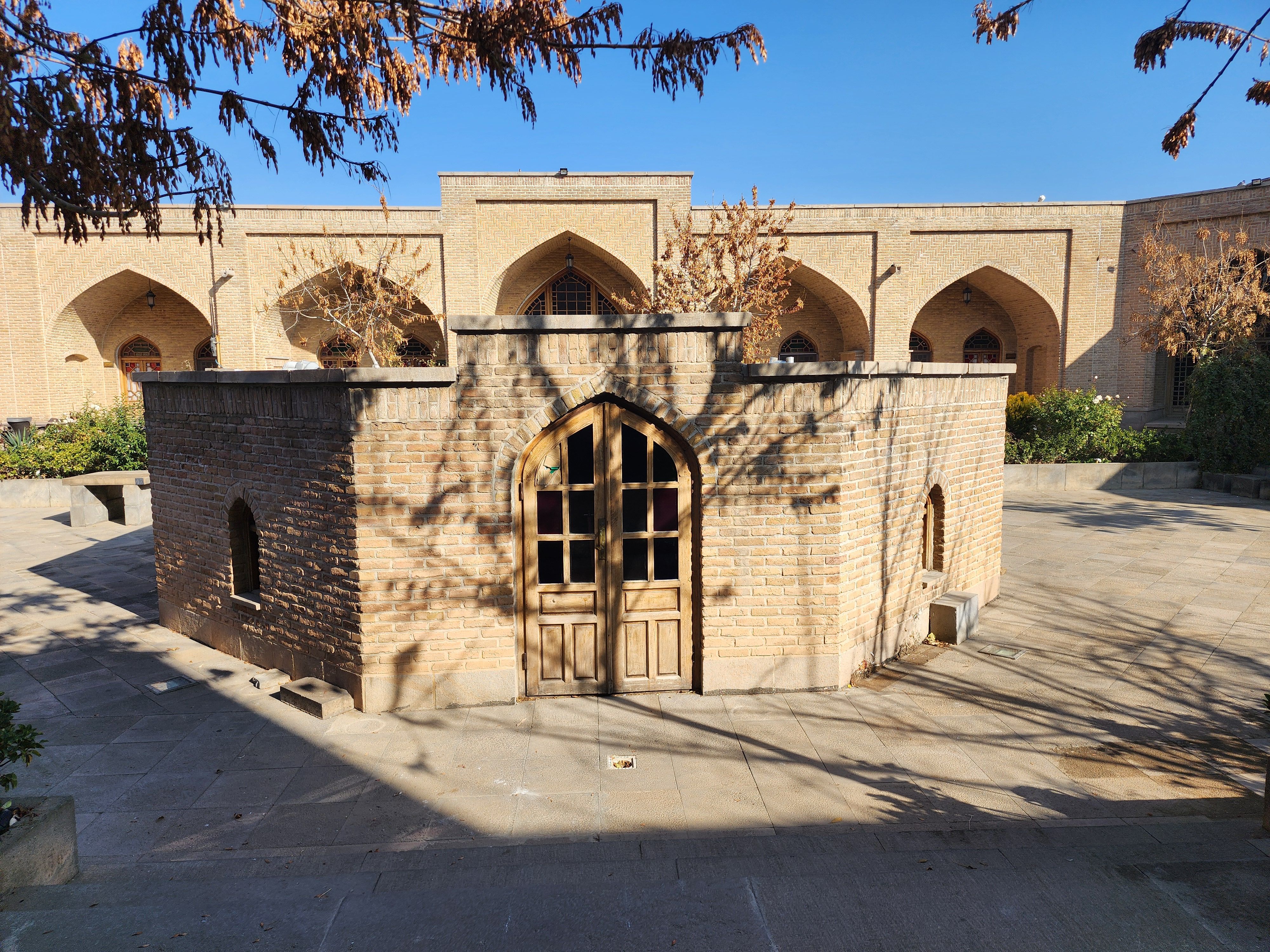 A sunny day in a historic courtyard with brick arches and dried trees casting shadows.
