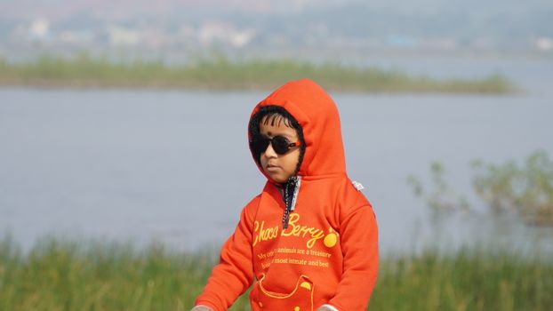 A young child wearing an orange hoodie stands near a peaceful lake in Jamshedpur.