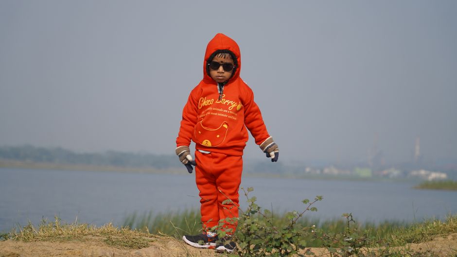 A young child wearing a bright red winter outfit stands by a riverbank in Jamshedpur, India.