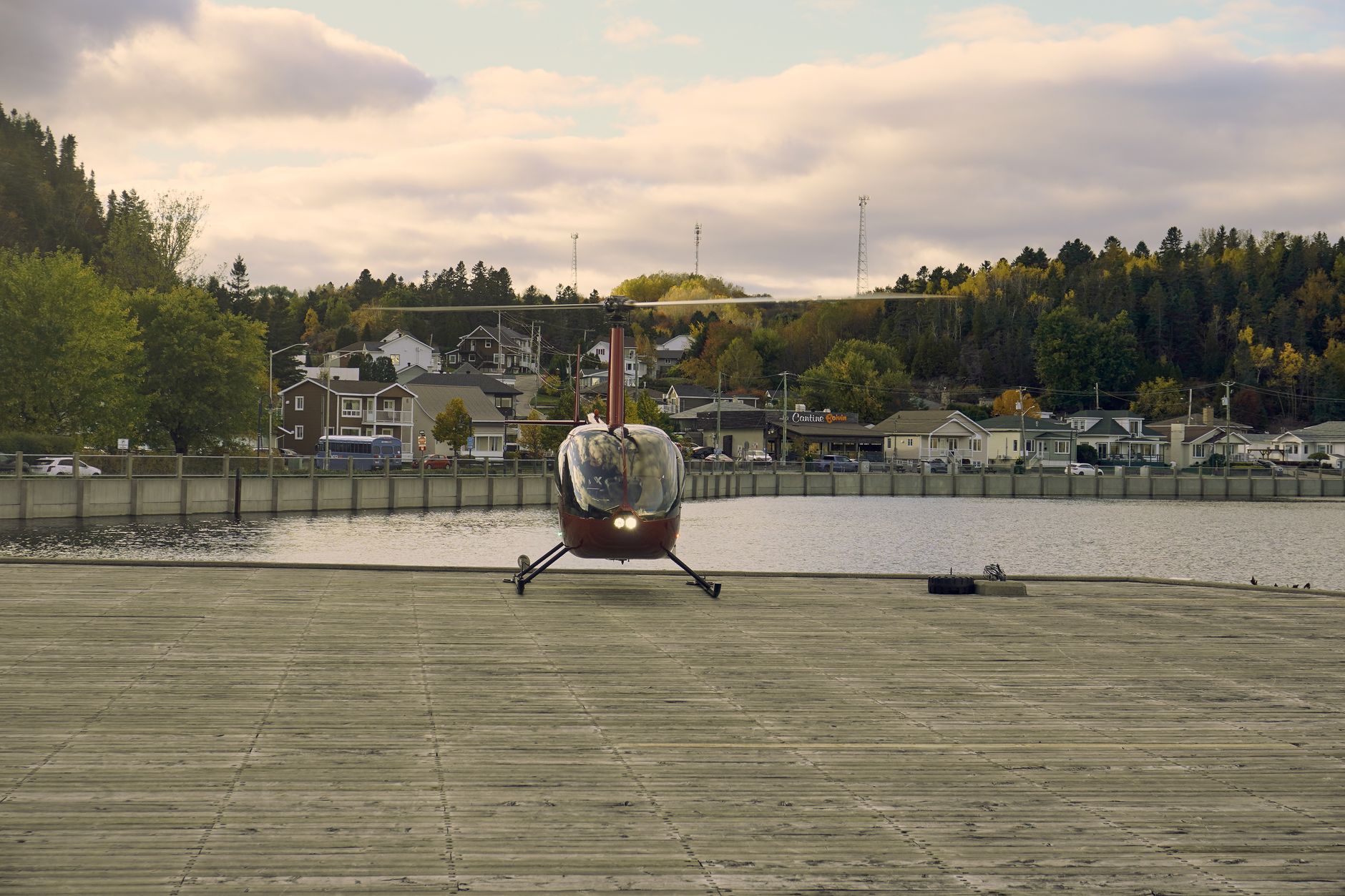 Red helicopter landing on waterfront platform amid scenic town and fall foliage.