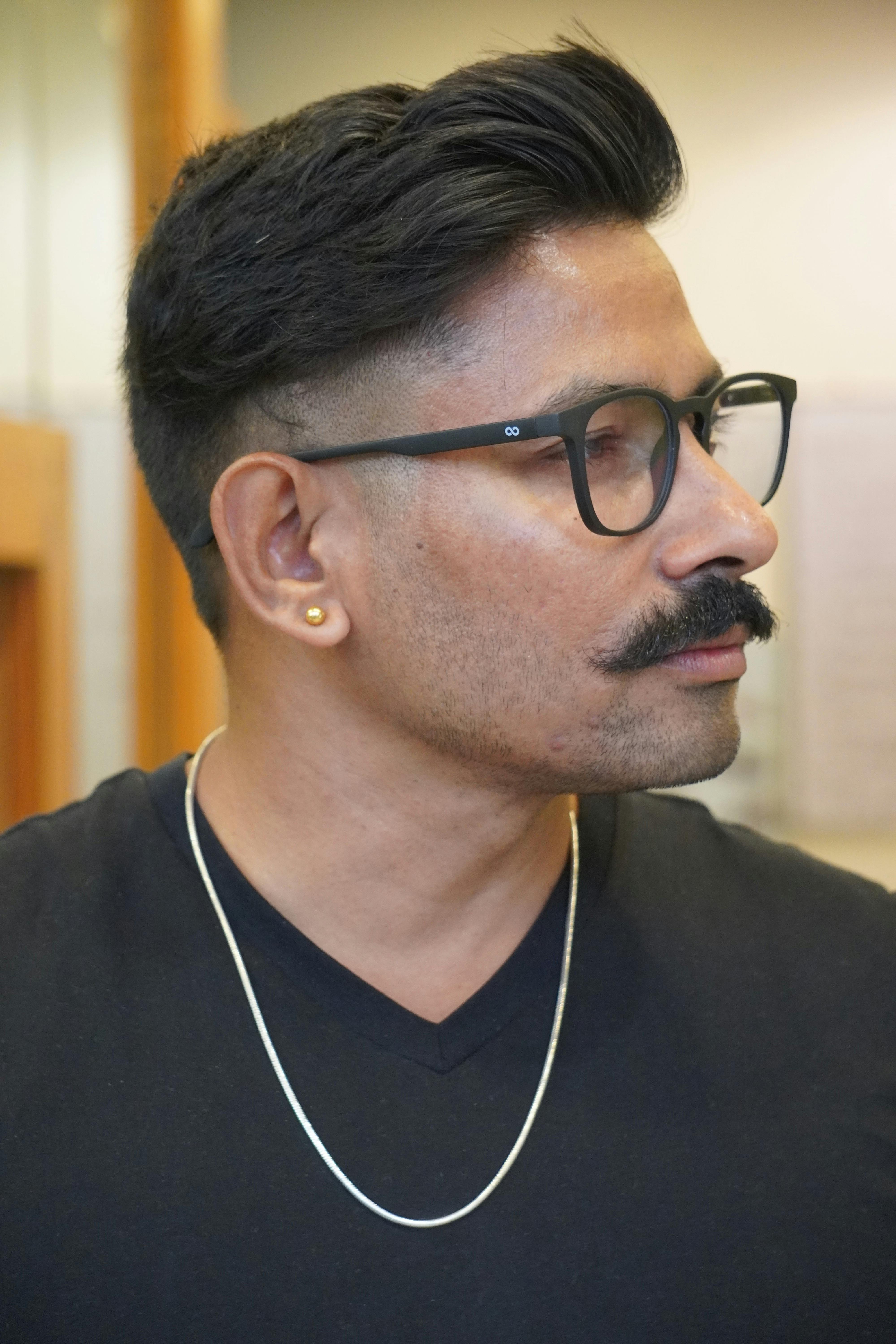 Close-up profile portrait of a stylish man with glasses and a mustache, wearing a black t-shirt indoors.