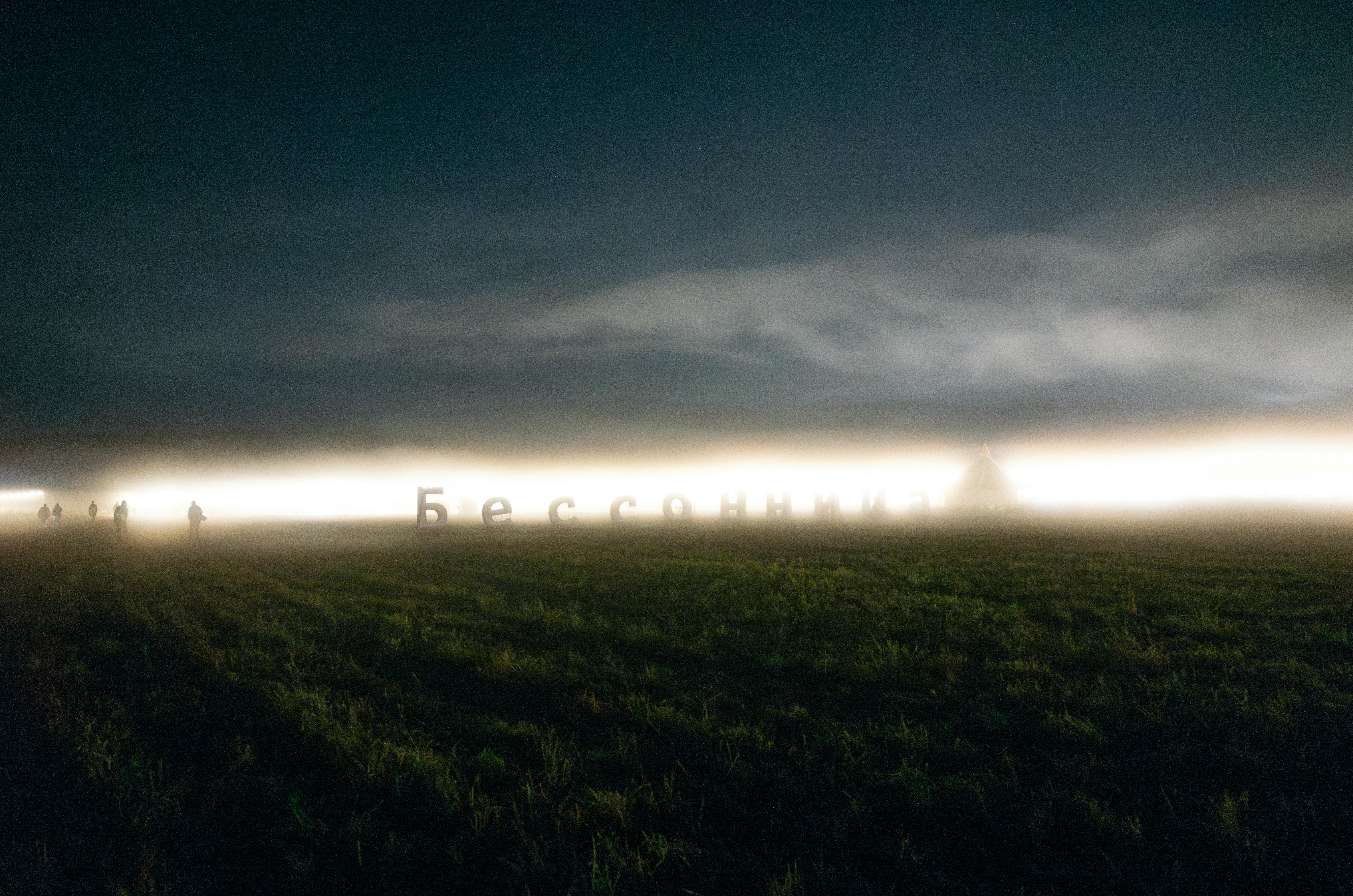 Foggy night landscape with a glowing light installation and silhouettes of people outdoors.