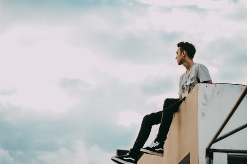 Free A young man sits on a building edge, looking afar into the sky. Stock Photo