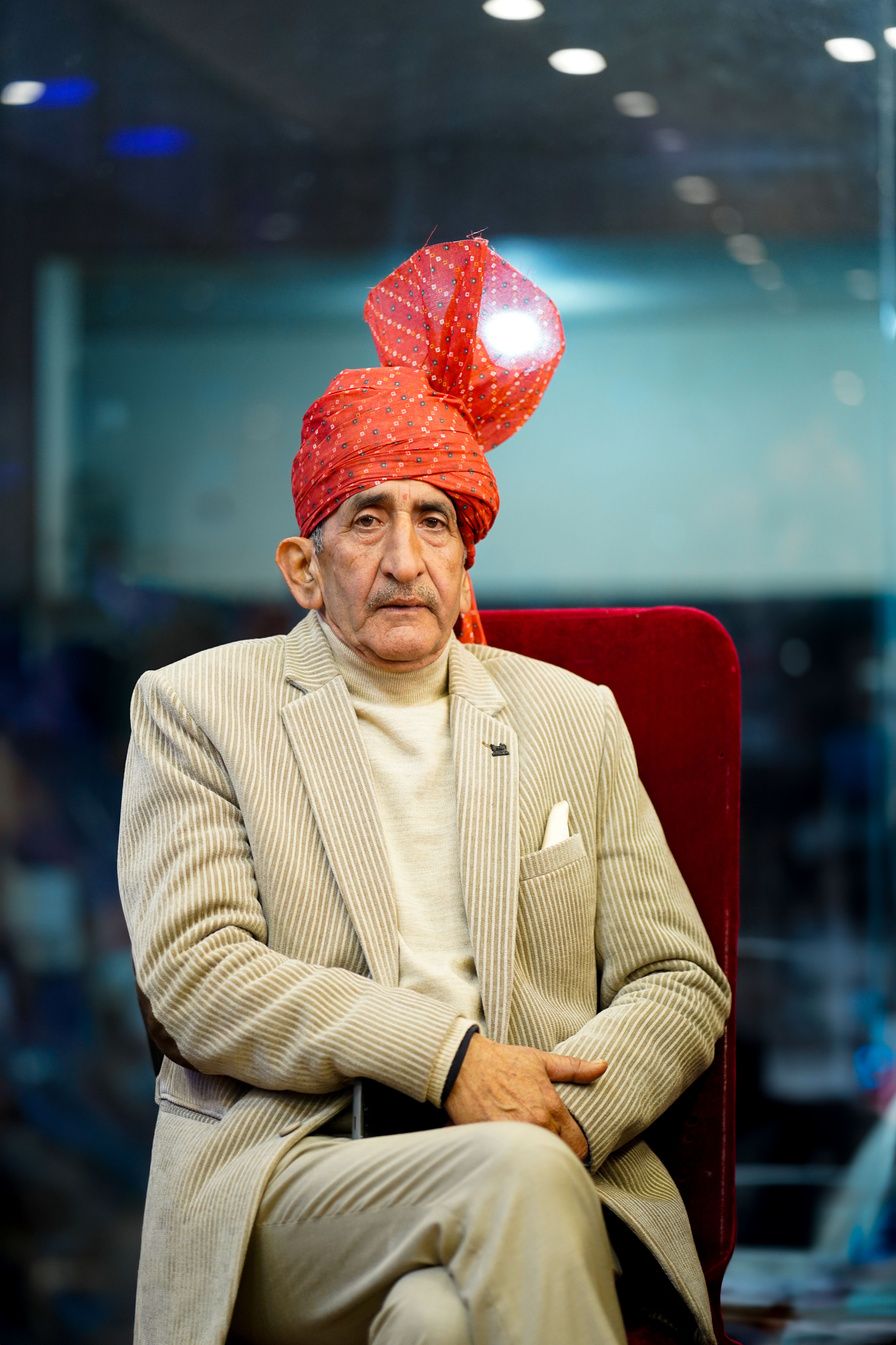 Senior man wearing a vibrant turban sitting indoors with a contemplative expression.