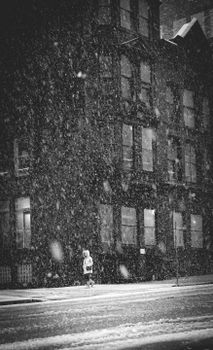 Black and white image of a person walking alone in a snowstorm on a city street at night.